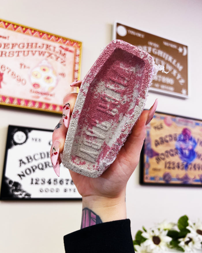 A hand with long pink manicured nails holds a small coffin-shaped graveyard bath bomb. The background shows a wall with framed Ouija boards and a blurred indoor setting.