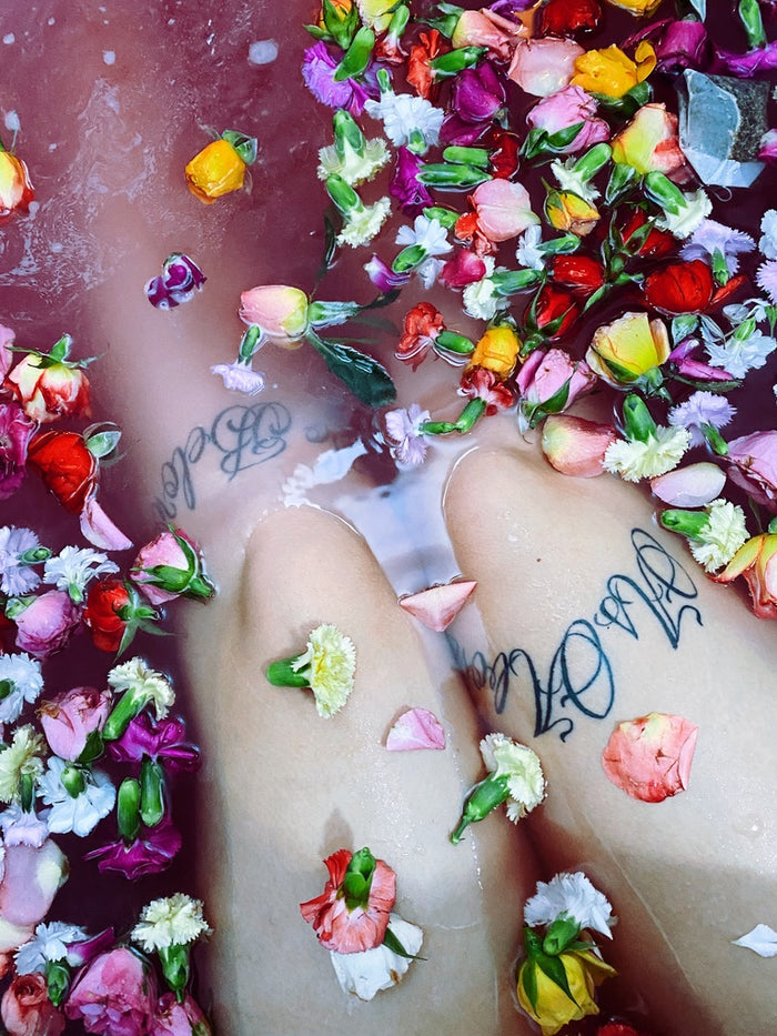 A close-up of a person's legs floating in pink bath water filled with colorful flower petals and small flowers. The person has black script tattoos on both legs.