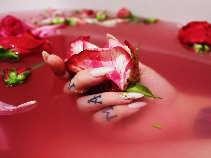 A hand with long white manicured nails and black letter tattoos holds a pink and white rose above a pink bath filled with floating rose petals.