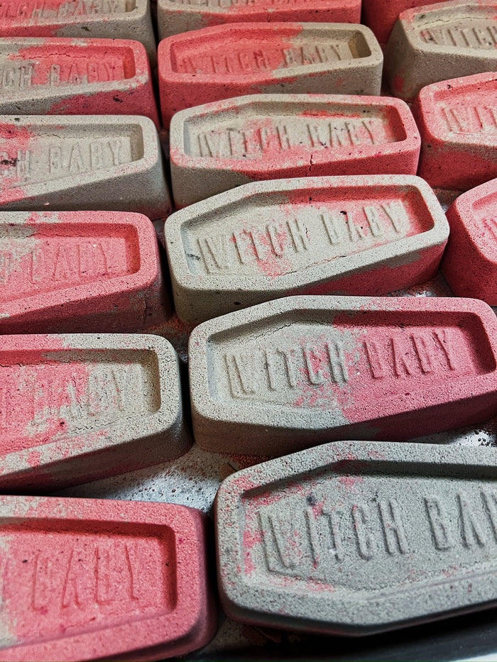 A close-up of multiple coffin-shaped bath bombs stamped with 'WITCH BABY' in raised letters. The soaps are shown in two colors: a light warm gray-beige and a dusty pink, with some soaps showing both colors and a few with red speckles.