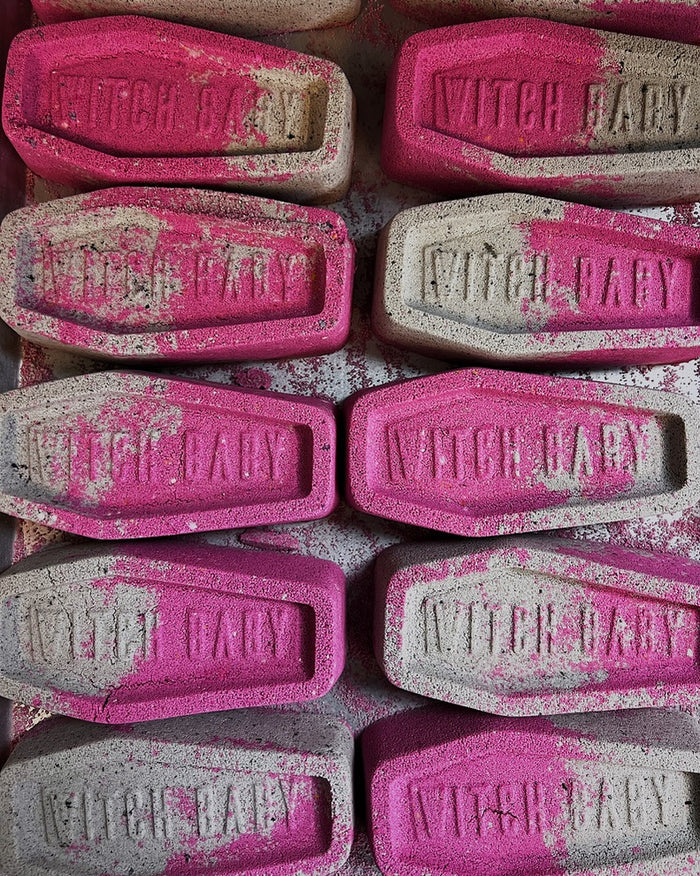 A close-up of multiple coffin-shaped bath bombs, featuring a textured pink and gray design with the words 'WITCH BABY' embossed on each piece.