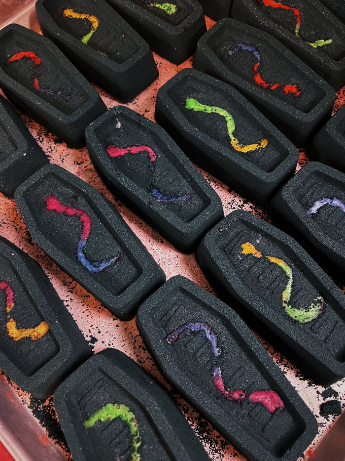 A close-up of multiple black coffin-shaped bath bombs arranged in rows on a metal tray. Each item has a small soapy gummy worm enmbedded in it in different colors including red, yellow, green, blue, and purple.