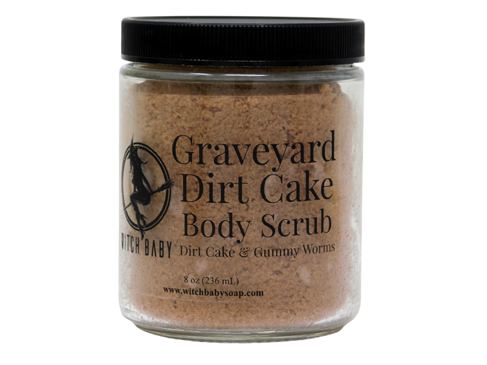 A clear plastic jar of Witch Baby Graveyard Dirt Cake Body Scrub with a black lid, containing a brownish-tan scrub, on a white background.