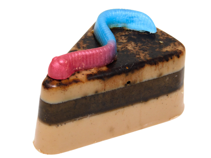A triangular slice-shaped chocolate confection soap styled like a slice of cake, with beige and dark brown layers, topped with a pink-and-blue gummy worm on a plain white background.