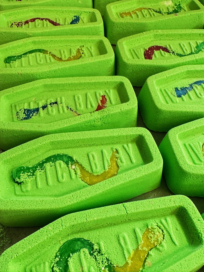 Bright green coffin shaped bath bombs with 'WITCH BABY' embossed on the top, each featuring colorful gummy worm soaps