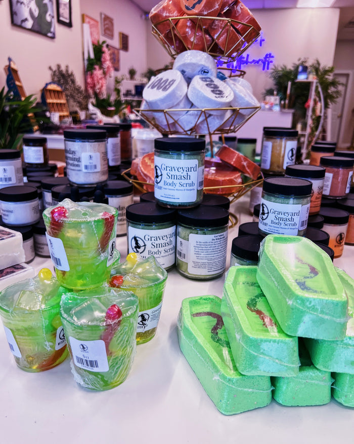 A display of bath products on a counter, including green translucent body scrubs in jars, stacked light green bars, and wrapped scrubs in a wire rack. The labels read 'Graveyard Smash' body scrub.