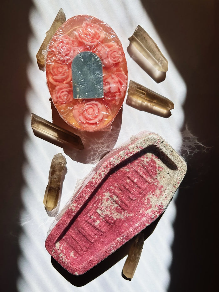 A Halloween-themed product display featuring a pink rose-patterned oval soap and a matching coffin-shaped mold, surrounded by raw crystal points on a dark background with dramatic shadow patterns.