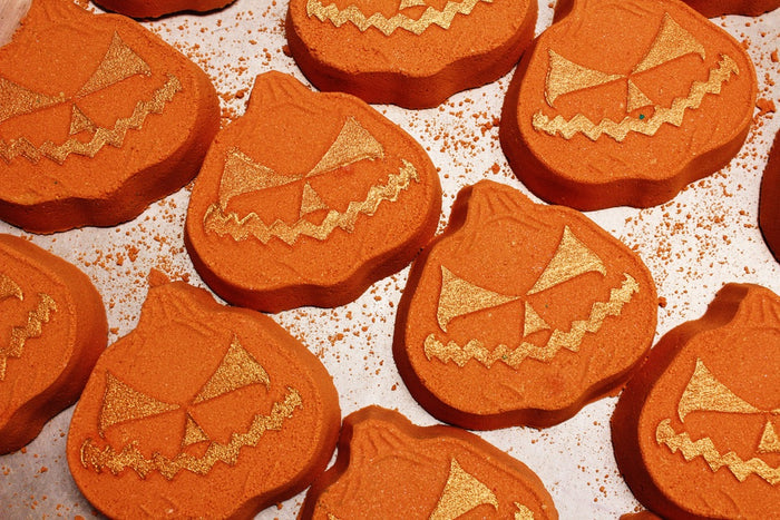 Orange pumpkin-shaped bath bombs with gold glitter jack-o'-lantern faces, arranged closely together on a white surface with scattered gold glitter.