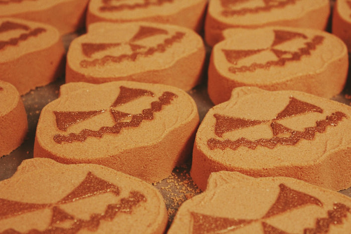 A close-up of pumpkin-shaped bath bombs with a carved jack-o'-lantern face, arranged closely together on a baking sheet.