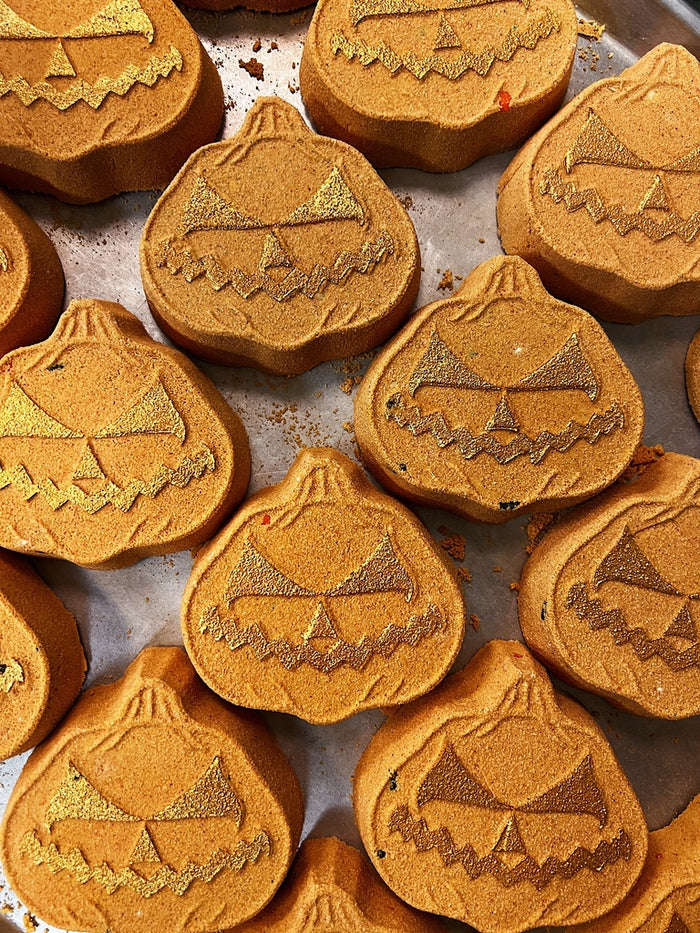 A close-up of multiple pumpkin-shaped bath bombs arranged on a baking sheet. The bath bombs are orange with a textured, crumb-like surface and are decorated with gold glitter accents in the carved facial features.