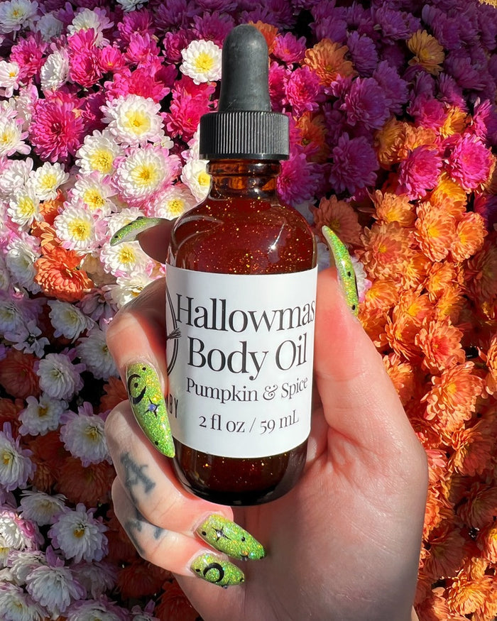 A hand with green glittery Halloween-themed nail art holds an amber glass dropper bottle of Halloween Body Oil. The background is a dense arrangement of colorful flowers in shades of pink, orange, and white.