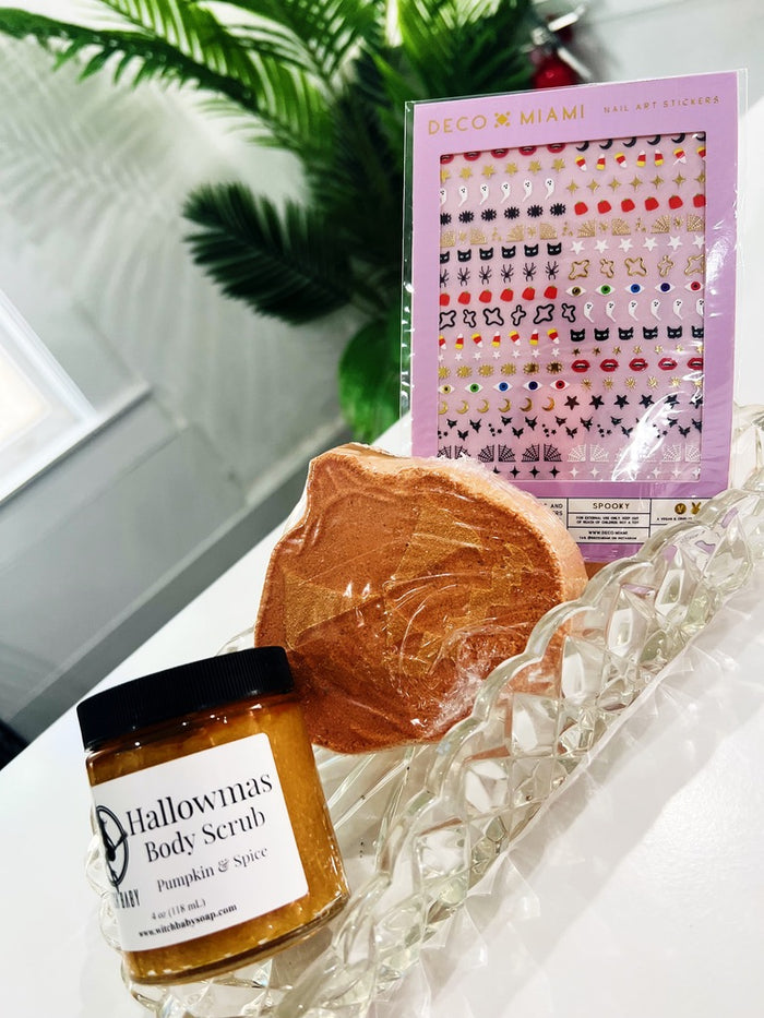 A styled product arrangement on a clear glass tray featuring a jar of Halloween-themed body scrub, a pumpkin bath bomb, and a package of Deco x Miami nail art stickers, with a window and green leaves in the background.