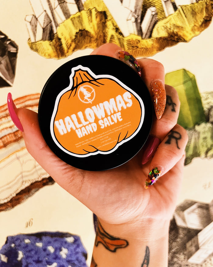 A hand with colorful glittery and painted nails holds a round black tin of 'Halloween Hand Salve' against a background of vintage-style illustrated cards.