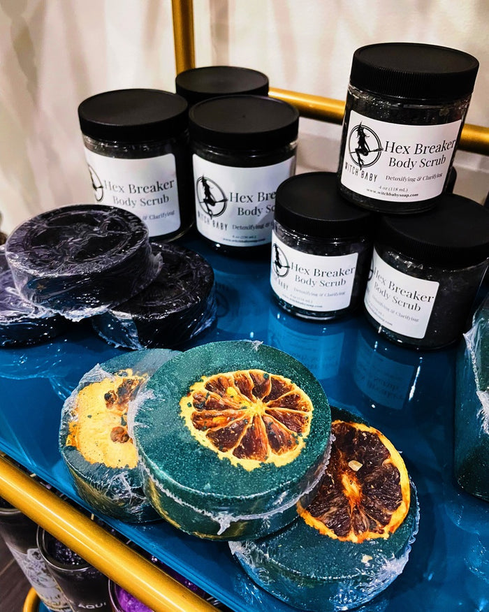 Several jars of Hex Breaker Body Scrub with black lids and white labels are arranged on a blue tray, alongside round dark scrub bars wrapped in plastic. The front bath bombs feature citrus slice designs, including dried lemon slices