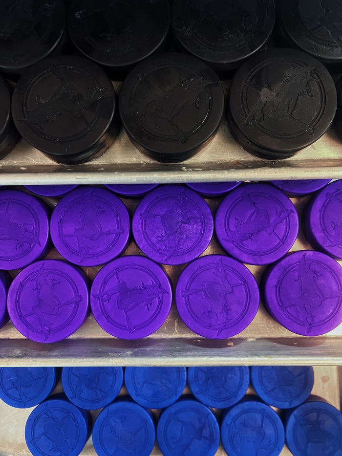 Three rows of round, witch baby soaps discs in black, purple, and blue, arranged on metal shelves. Each disc has an embossed witch design in the center.