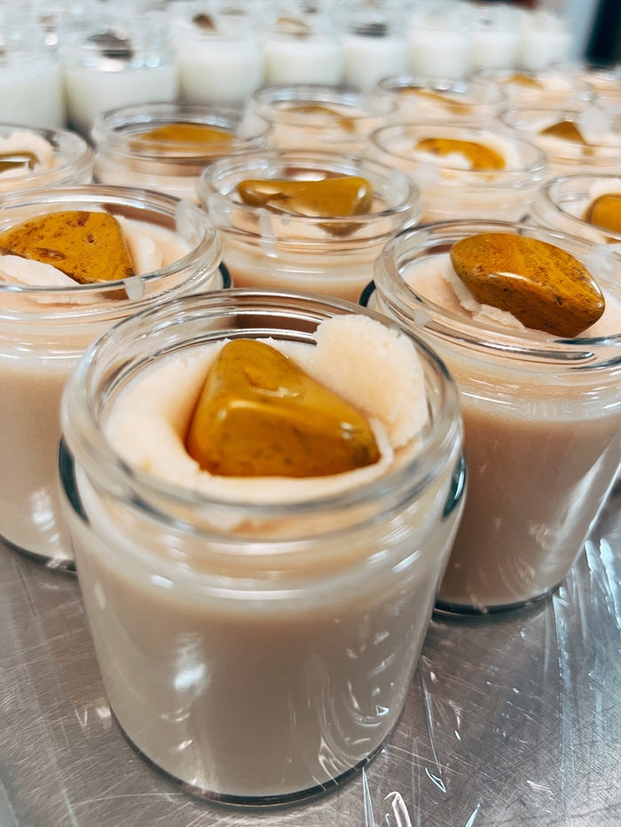 Multiple small clear glass jars filled with a honey colored body butter, each topped with a glossy amber-brown piece yellow jasper, arranged on a metallic surface.