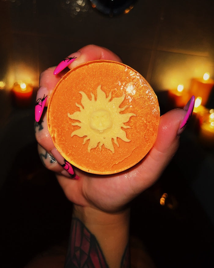 A hand with bright pink nails holds a round orange soap disc carved with a yellow sun design in the center, set against a dark background with blurred warm candlelight.