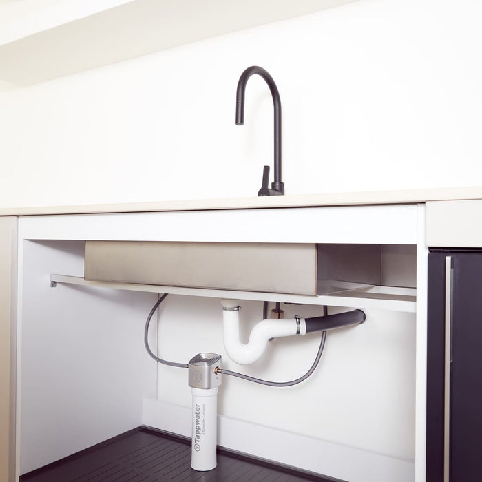Un moderno lavello da cucina con rubinetto nero è installato sopra un mobile. All'interno sono visibili i tubi e un filtro in linea Tappwater SMR. Lo spazio è pulito e minimalista.