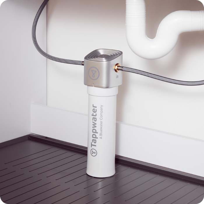A Tappwater Inline Filter SMR with grey pipes is installed under a kitchen sink, connected to the plumbing. The dark, grooved cabinet floor is visible beneath the unit.