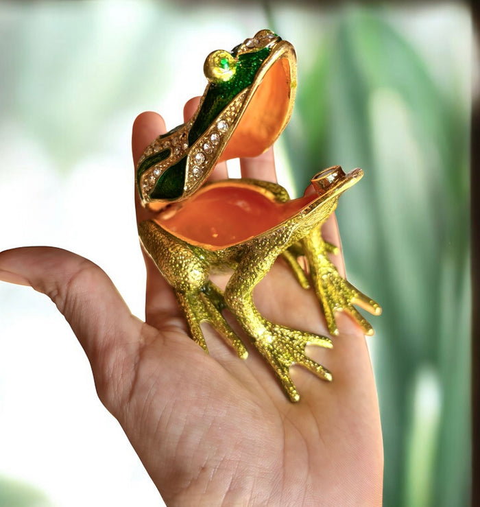 Hand holding open jeweled green enamel frog trinket box showing warm terracotta satin interior, green botanical background
