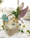 Jeweled hummingbird trinket box with green rhinestone body and lavender wings perched on cream enamel box, displayed on a floral-patterned cloth with soft pink background