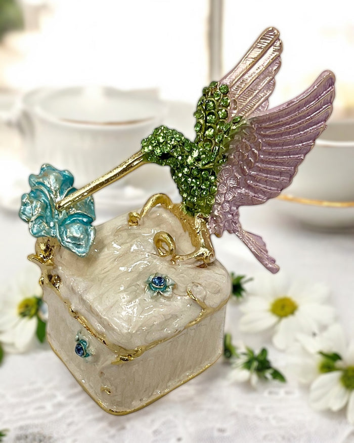 Jeweled hummingbird trinket box with green rhinestone body and lavender wings perched on cream enamel box, displayed on a floral-patterned cloth with soft pink background
