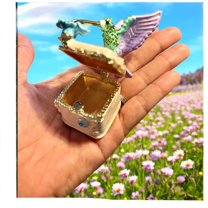 Jeweled hummingbird trinket box displayed on top of a gold brocade gift box, white background, showing full hummingbird and blue enamel flower detail