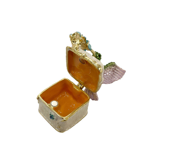 Side view of jeweled hummingbird trinket box with green rhinestone body, lavender wings, and cream enamel base on white background