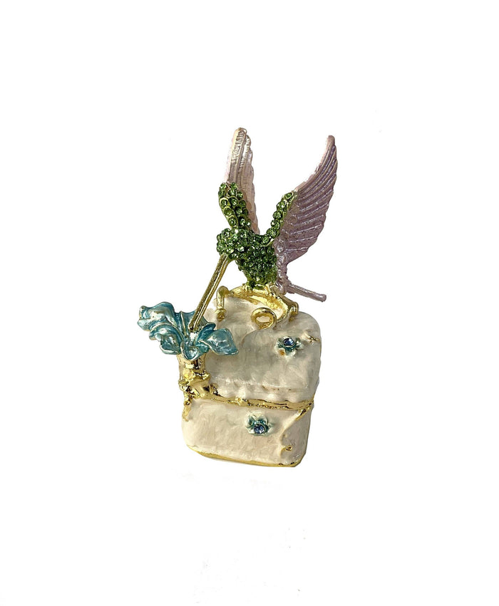 Front view of jeweled hummingbird trinket box with green rhinestone hummingbird, lavender enamel wings, and blue flower on cream enamel box, white background