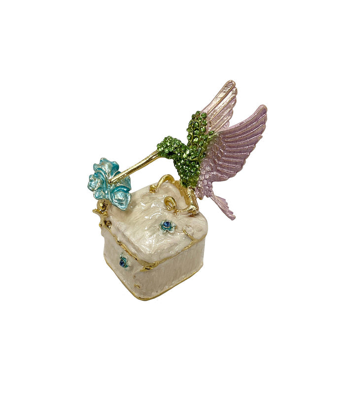 Three-quarter view of jeweled hummingbird trinket box closed, showing cream enamel body with gold trim and blue floral rhinestone accents, white background