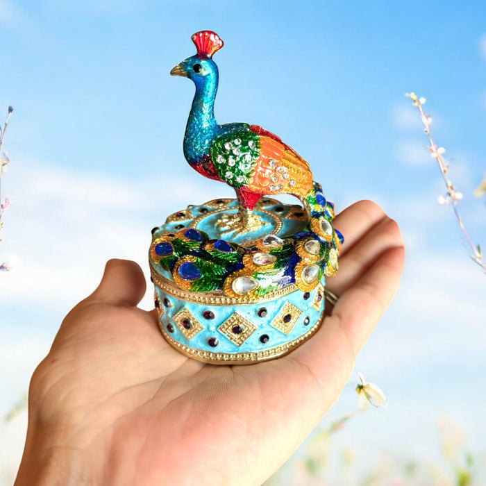 Hand holding multicolor enamel peacock trinket box with teal and green peacock sculpture perched on a sky blue rhinestone-covered round box, blue sky background