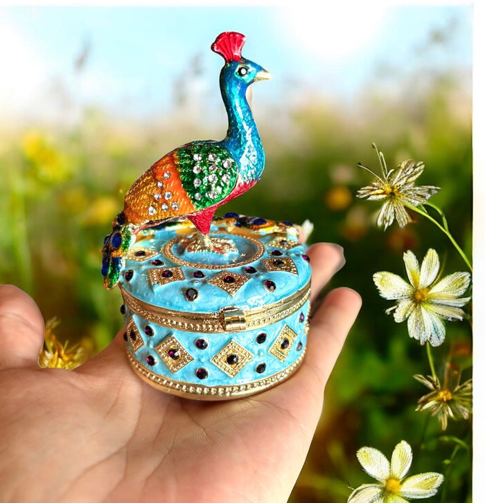 Hand holding jeweled peacock trinket box with multicolor enamel peacock and rhinestone feather detailing on sky blue box, sunny wildflower field background