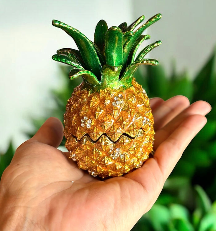 Hand holding gold enamel pineapple trinket box with rhinestone accents and green sculpted crown leaves, soft green tropical plant background
