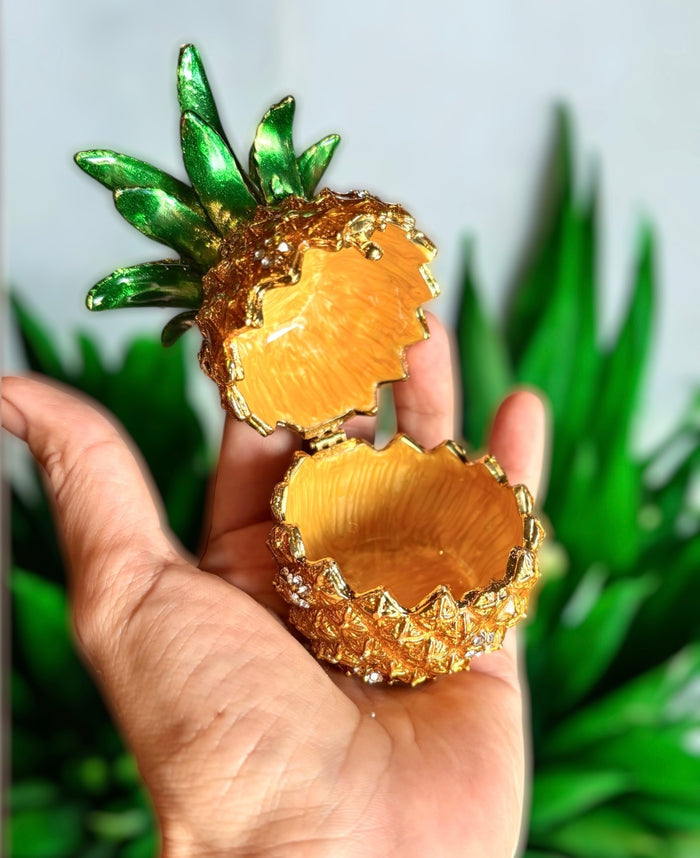 Hand holding open gold enamel pineapple trinket box showing warm golden interior, with green tropical plant background