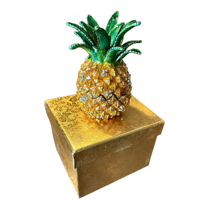 Gold enamel pineapple trinket box with rhinestone accents and green crown leaves displayed on top of gold brocade gift box, white background