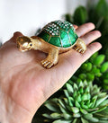 Hand holding gold tone jeweled tortoise trinket box with emerald green rhinestone shell, surrounded by green succulent plants
