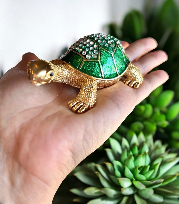 Hand holding gold tone jeweled tortoise trinket box with emerald green rhinestone shell, surrounded by green succulent plants
