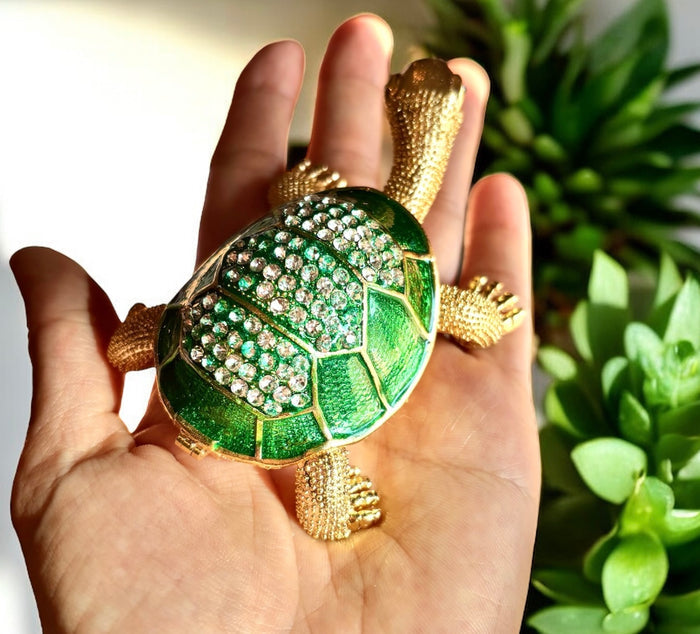 Top-down view of jeweled tortoise trinket box with emerald green enamel shell fully covered in clear rhinestones, held in an open hand with succulents in background