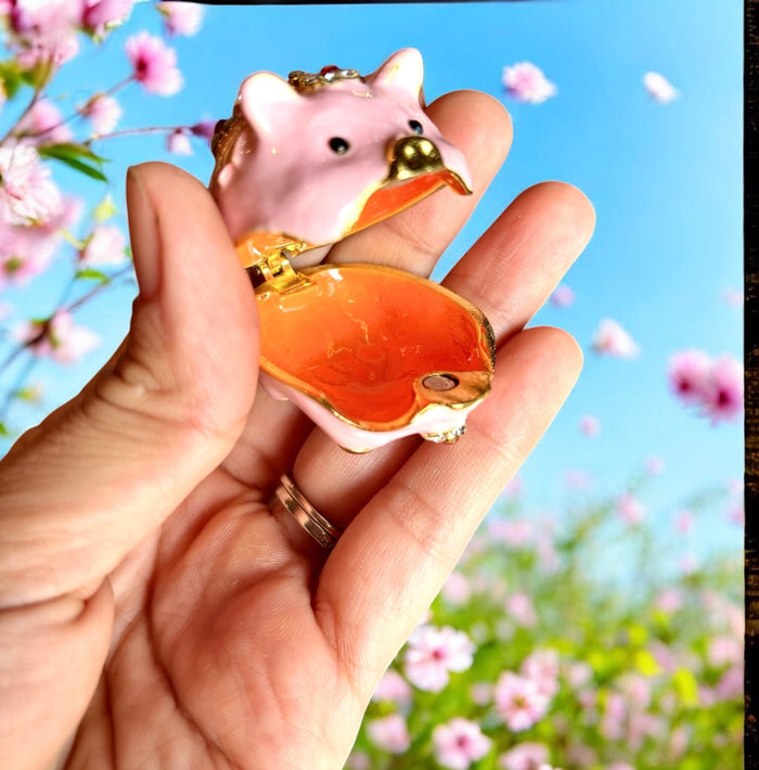 Pink enamel jeweled pig trinket box displayed on top of a gold brocade gift box, white background