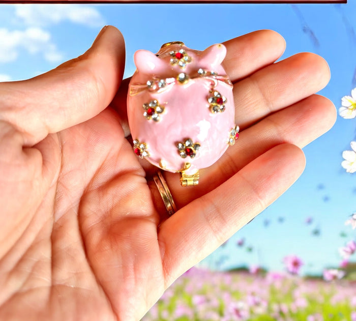 Pink enamel pig trinket box nestled inside open gold brocade gift box with warm orange satin lining, white background