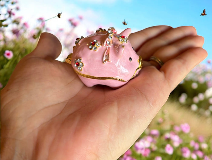 Hand holding pink enamel pig trinket box with rhinestone flower clusters, pink cosmos flower field background