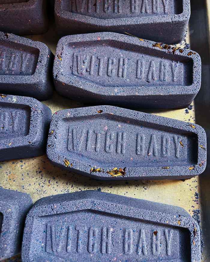 A close-up of several dark lavender gray, coffin-shaped bath bombs embossed with the brand name 'WITCH BABY' on a speckled beige surface.