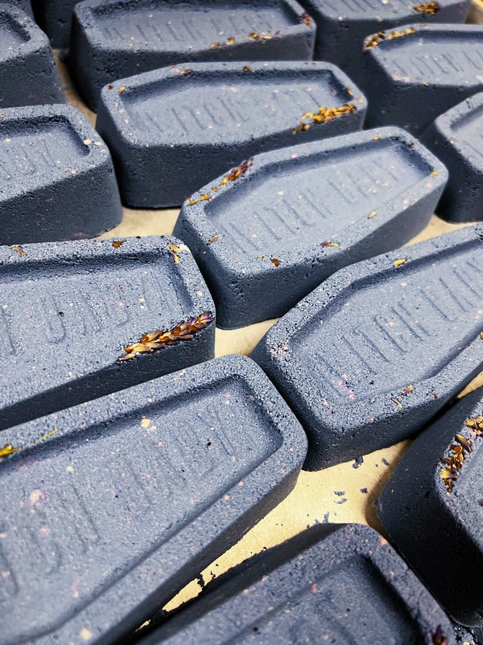 A close-up view of multiple lavender coffin bath bombs, arranged on a light beige surface. The bath bombs have a textured matte finish with small flecks and dried botanical pieces embedded in them.