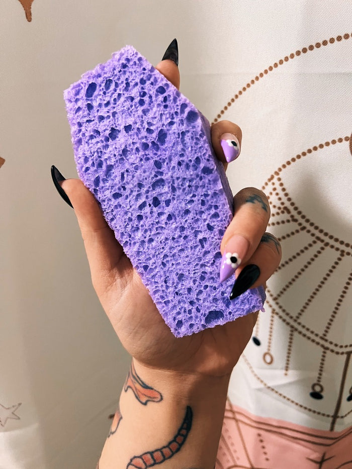 A hand with black and purple nail art holds a purple porous coffin shaped sponge against a patterned curtain background.