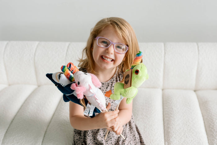 Smiling young girl with glasses holding a bundle of four Lollyplush animal toys including a shark, unicorn, sloth, and dinosaur, seated on a white couch