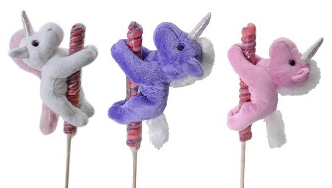 Three Lollyplush unicorn plush toys in white, purple, and pink each hugging a rainbow lollipop on a wooden stick, white background