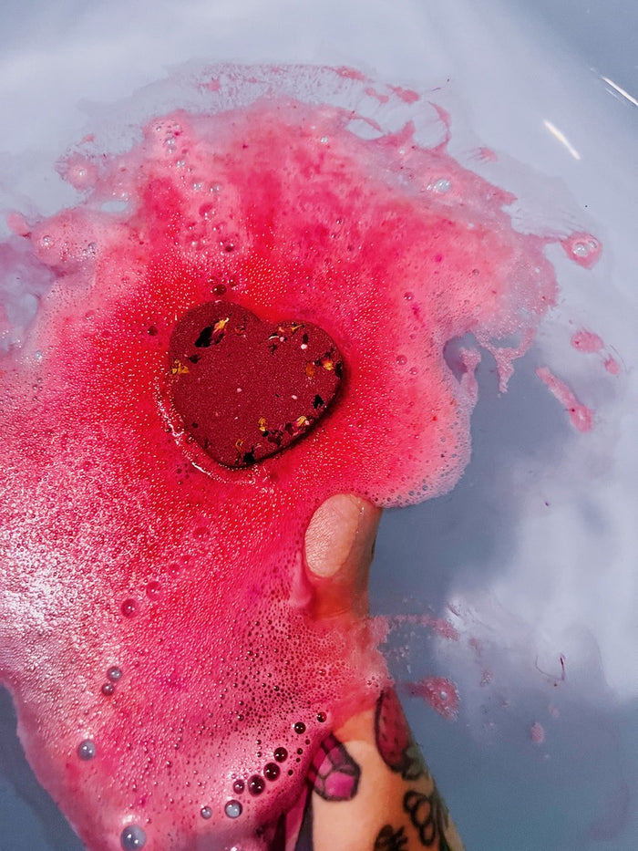 A close-up of a hand dropping a heart-shaped bath bomb into soapy pink water, creating a burst of pink foam and bubbles.