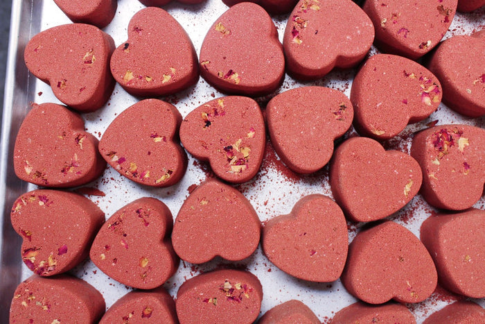 Heart-shaped bath bombs in a dusty rose-red color, scattered on a white tray with small dried flower petals visible inside.