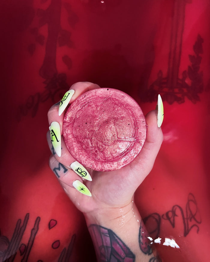 A hand with neon yellow and green manicured nails holds a round pink soap against a dark pink bath