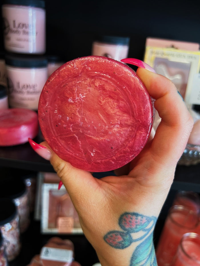 A hand with pink manicured nails and a strawberry tattoo holds a round pink soap in front of a dark shelf filled with other products.